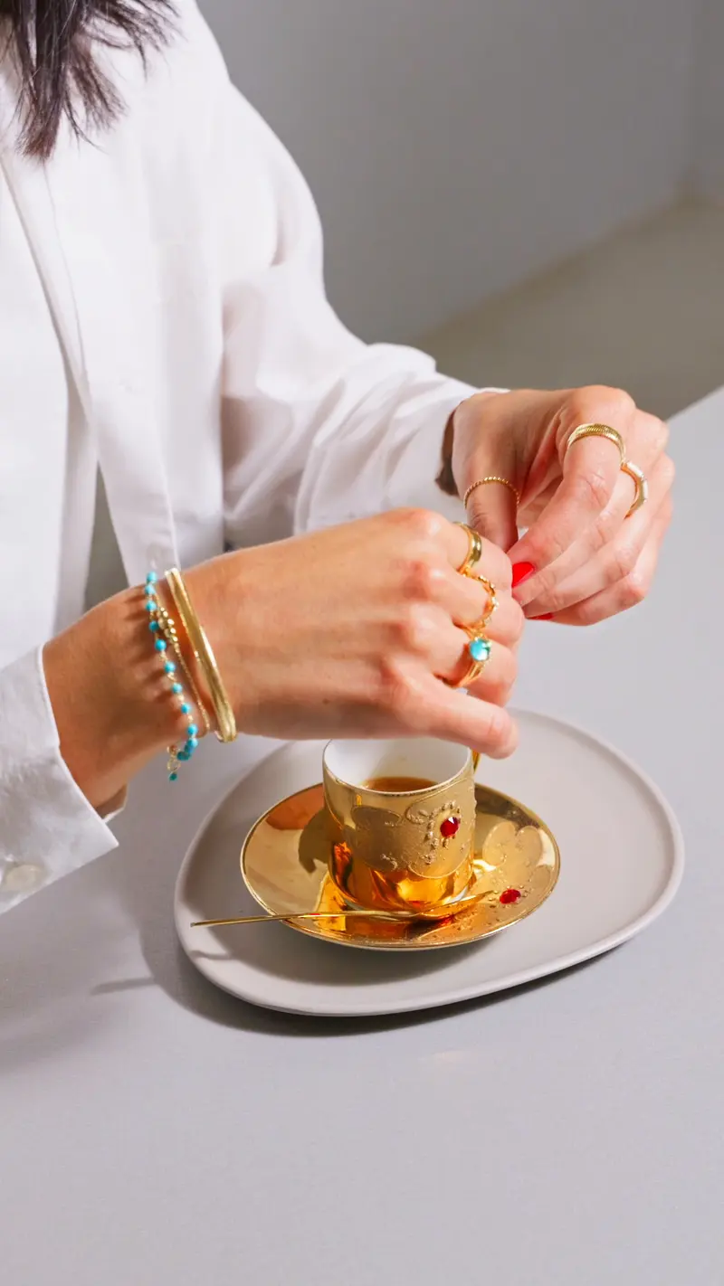 Mani con anelli e bracciali che sollevano una tazzina dorata dal piattino