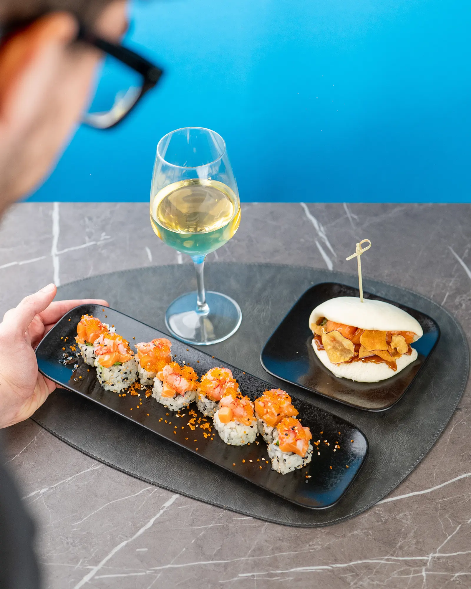 Vassoio di sushi e bao accanto a un calice, fondo azzurro