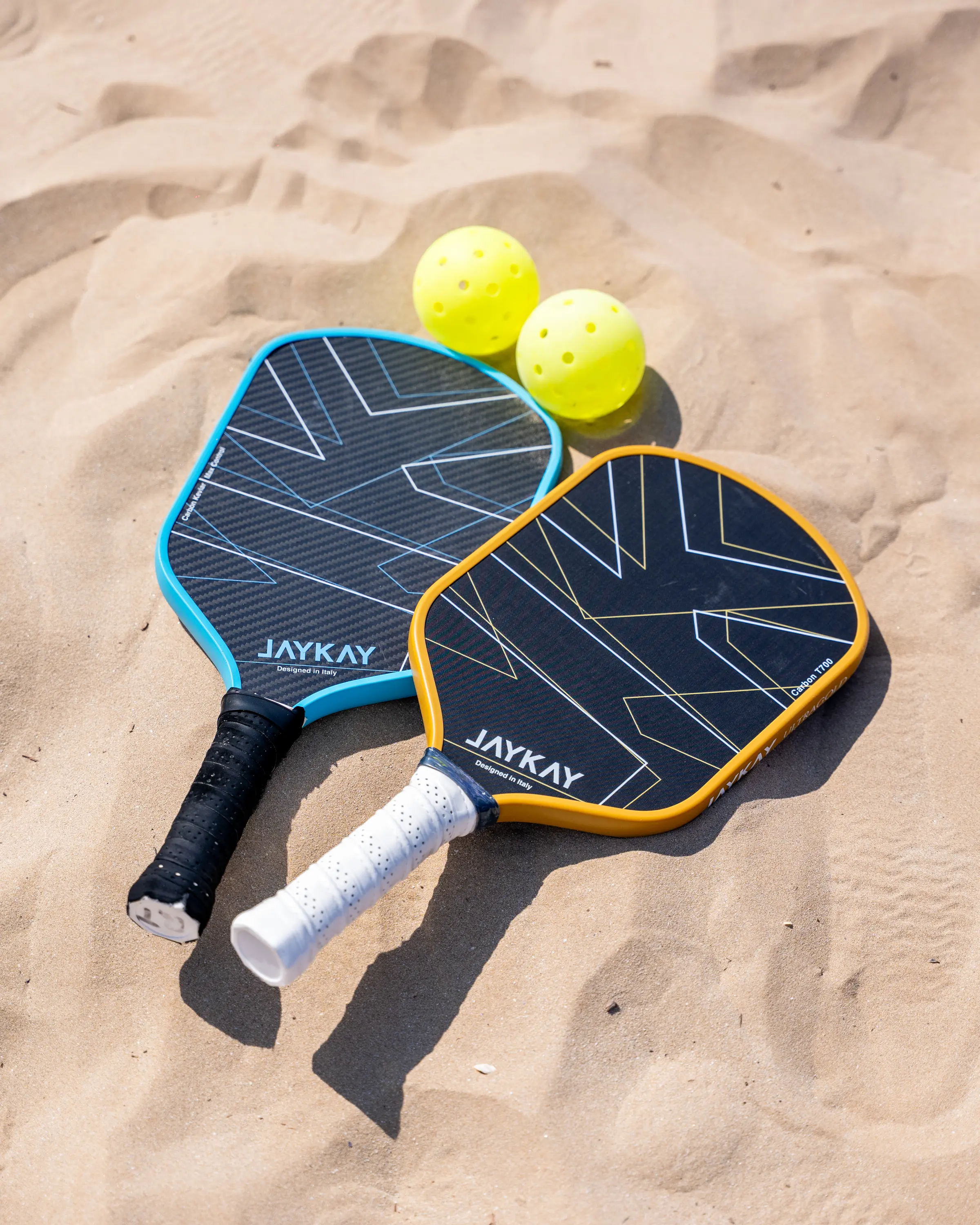 Due racchette da pickleball JAYKAY appoggiate sulla sabbia accanto a due palline