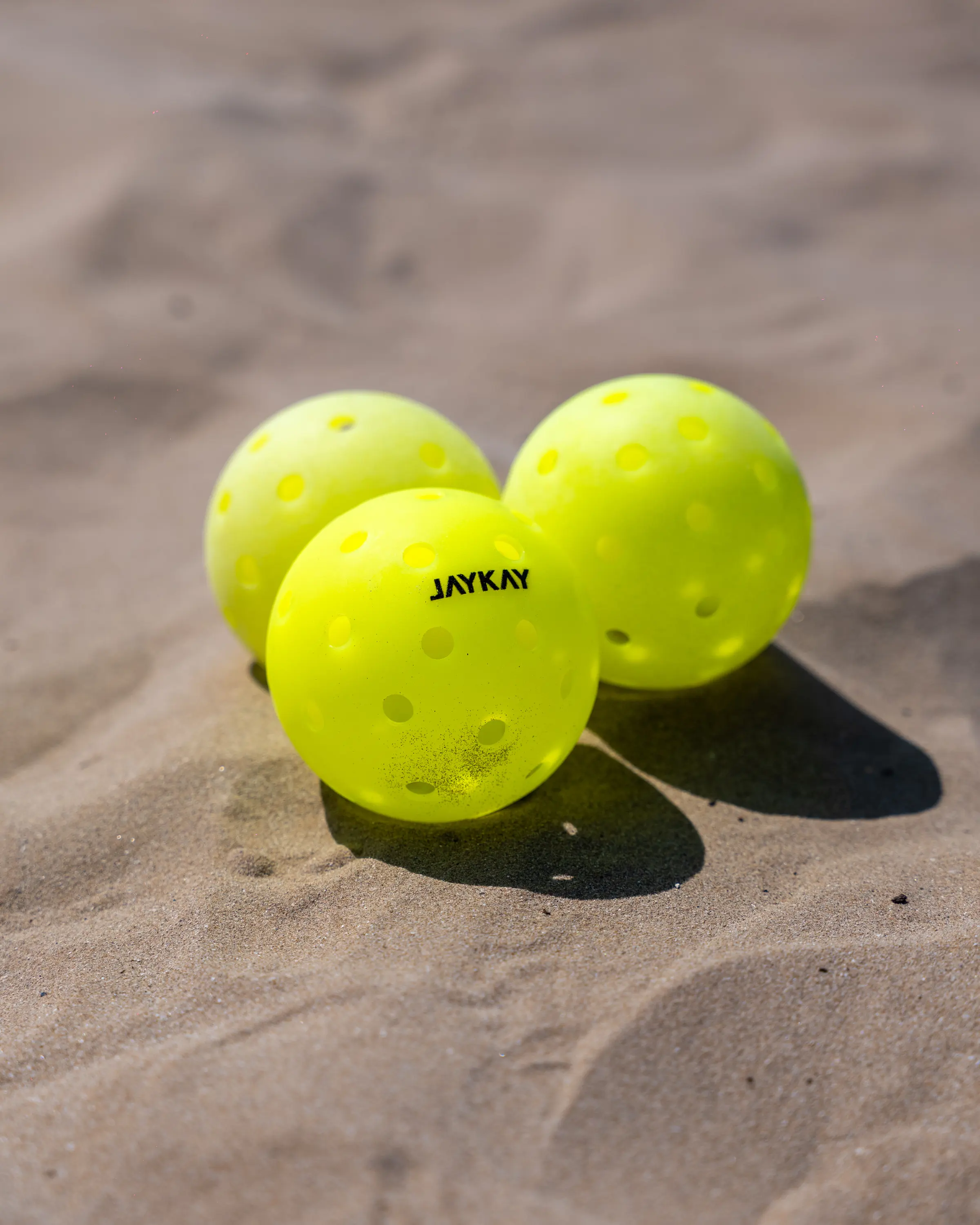 Tre palline gialle da pickleball sulla sabbia, una con il marchio JAYKAY