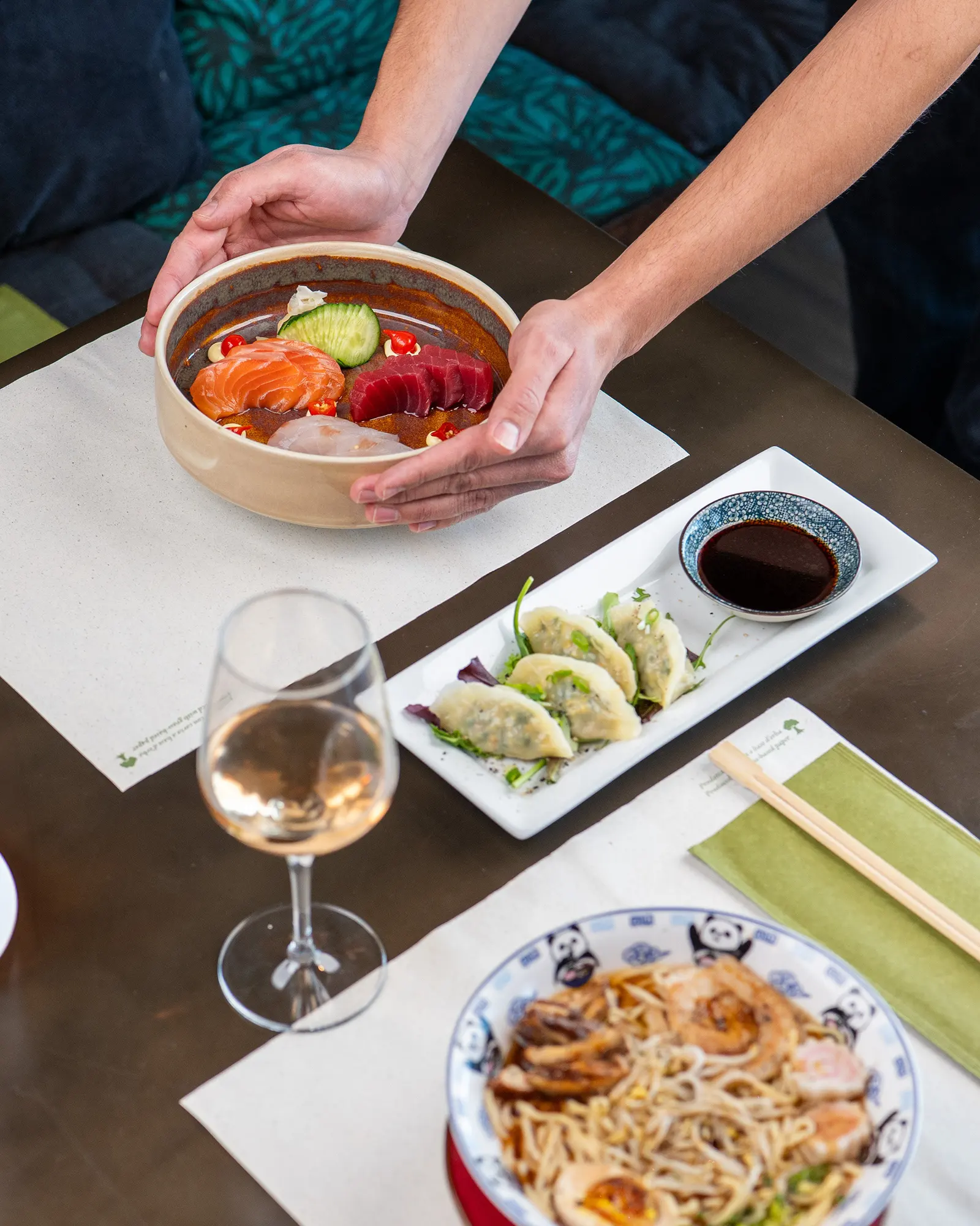 Le mani posano una ciotola di sashimi sul tavolo, accanto a gyoza e ramen