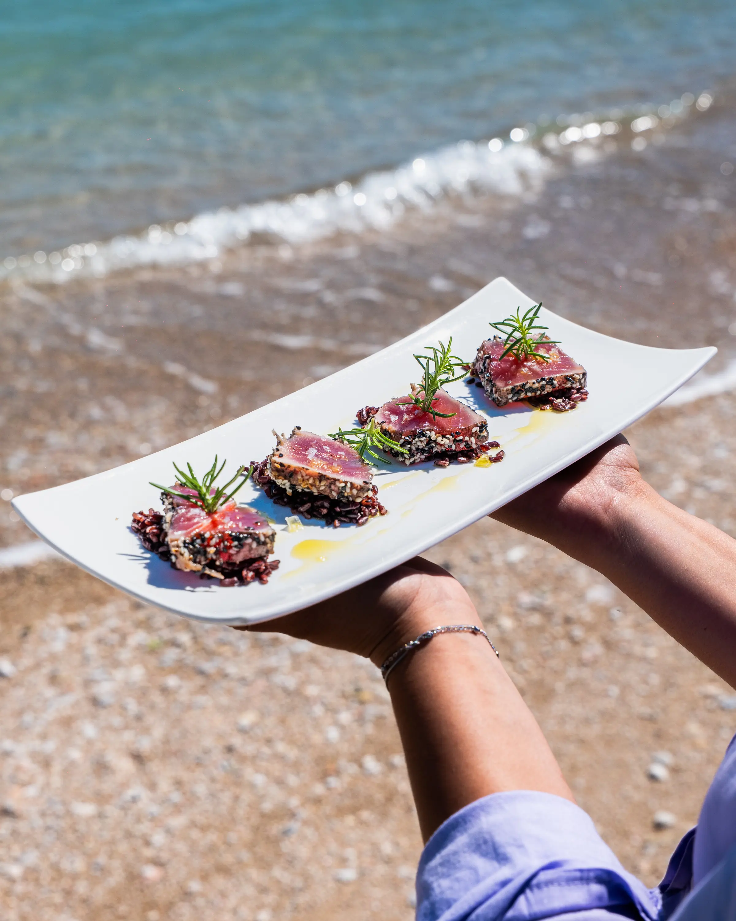 Piatto di crudi di pesce tenuto sulla battigia, con l’onda alle spalle
